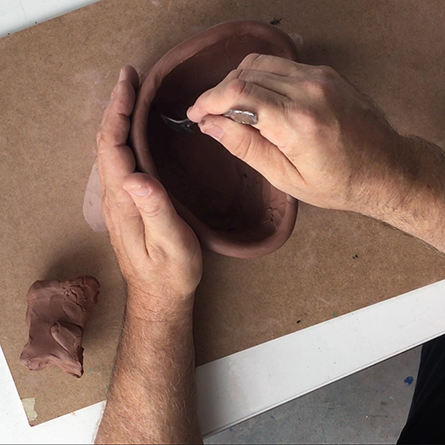 Two hands sculpting a clay pot with a spoon